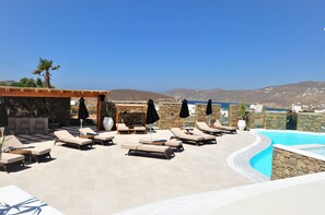 Zona de piscina en la azotea con sillones de descanso y sombrillas, un bar y vistas al mar y colinas.