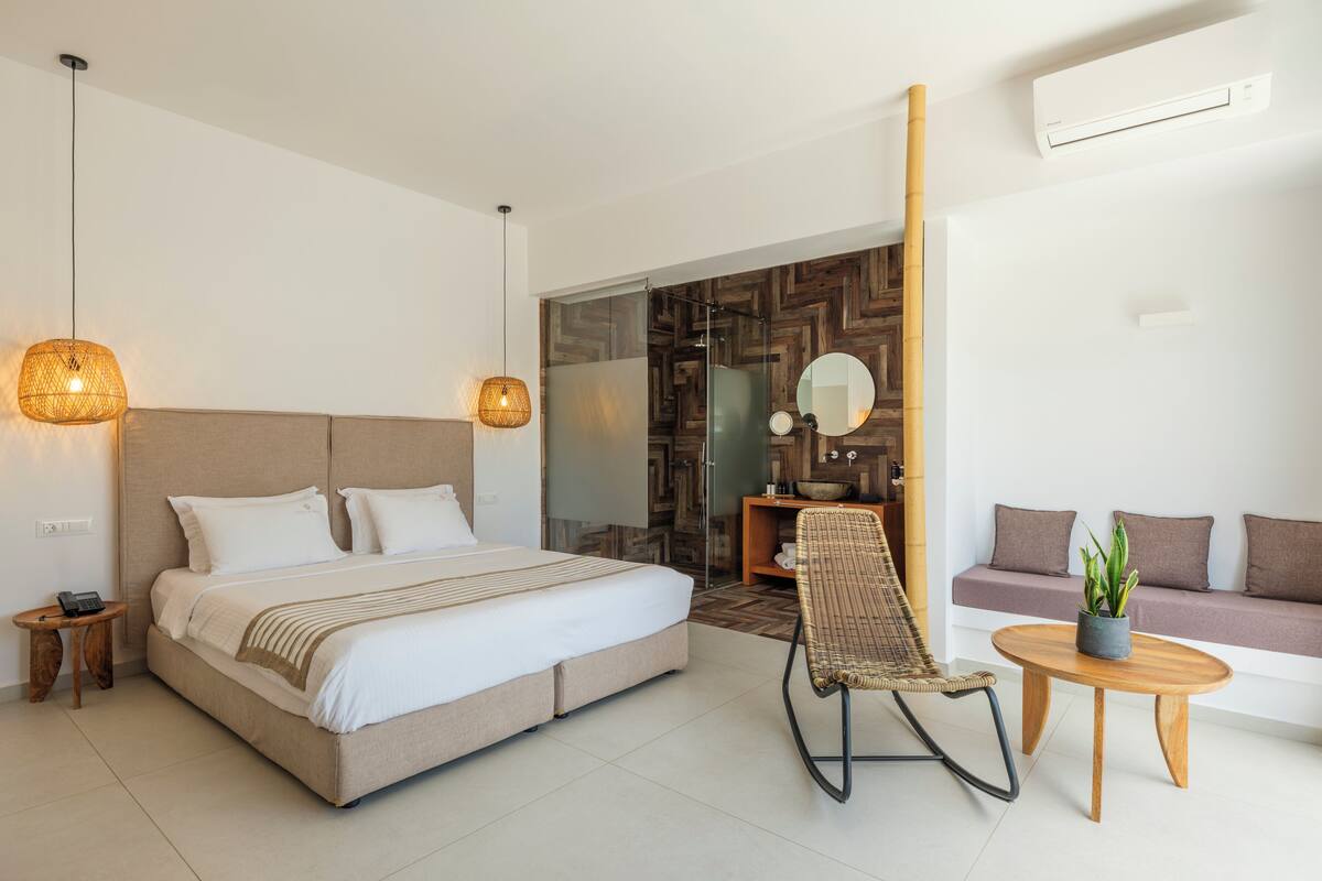 A modern hotel room with a large bed, a seating area with a sofa and a chair, a small table with a plant, and a bathroom visible through a glass door.