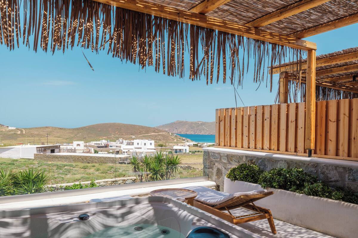 A hot tub with wooden loungers, a stone wall, and a view of the sea and buildings in the background.