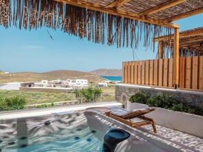 Un jacuzzi con tumbonas de madera, una pared de piedra y vista al mar y edificios en el fondo.