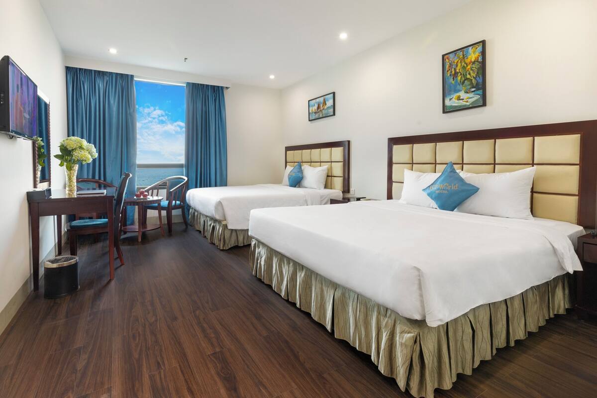 A hotel room with two beds, a desk, a chair, a TV, and a large window with curtains.