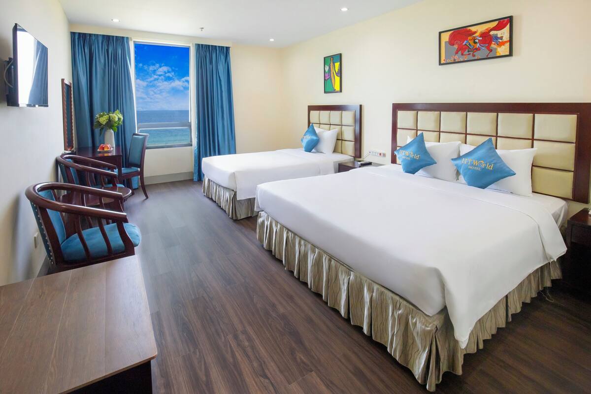 A hotel room with two beds, a desk, and a chair. There is a window with curtains, a painting on the wall, and a view of the ocean.