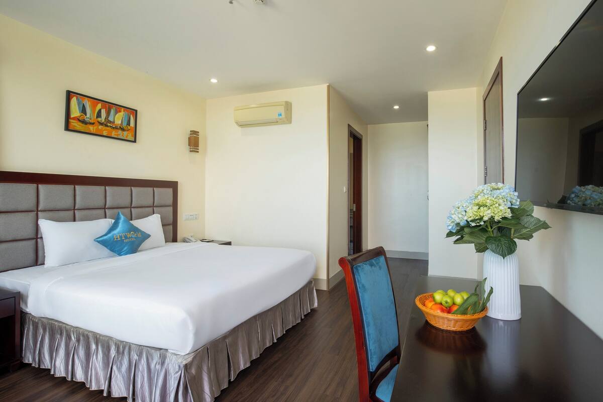 A hotel room with a large bed, a headboard, a painting, an air conditioning unit, a mirror, a vase with flowers, and a fruit basket.