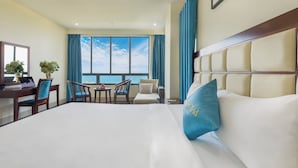 A hotel room with a large bed, a desk with a chair, a small table with a chair, and a view of the sea through large windows. - Sea Front Hotel (Da Nang)