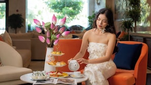 A woman in a white dress seated at a table with a teapot, a tray of pastries, and a vase of pink flowers. - Sea Front Hotel (Da Nang)