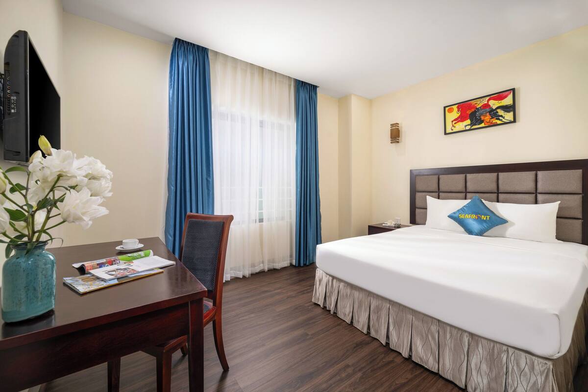 A hotel room with a bed, a desk with a vase of flowers, a chair, a television, and a painting on the wall.