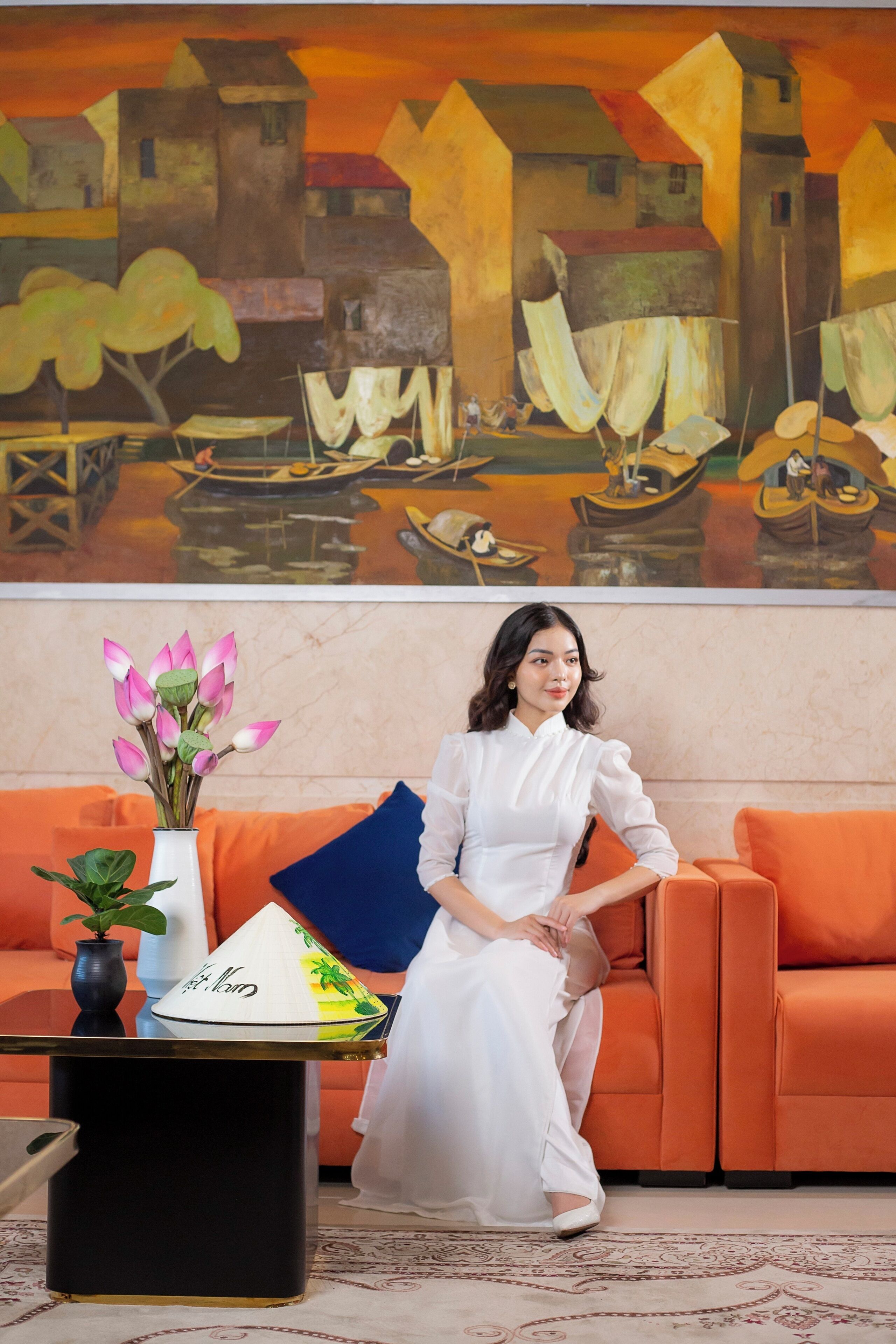 A woman seated on an orange sofa with a white vase of flowers and a decorative cone on a black table in front of her.