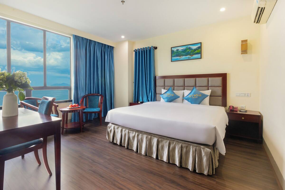 A hotel room with a large bed, a dining table, chairs, a view of the sea, and a painting on the wall.