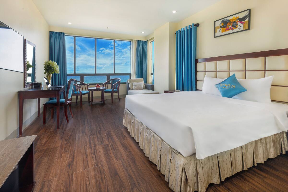 A hotel room with a large bed, a desk with a laptop, a chair, and a view of the sea through large windows.