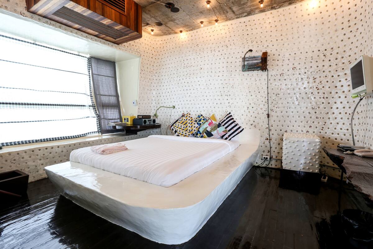 A room with a large bed, a window with blinds, a radiator, and a wall with a polka dot pattern.