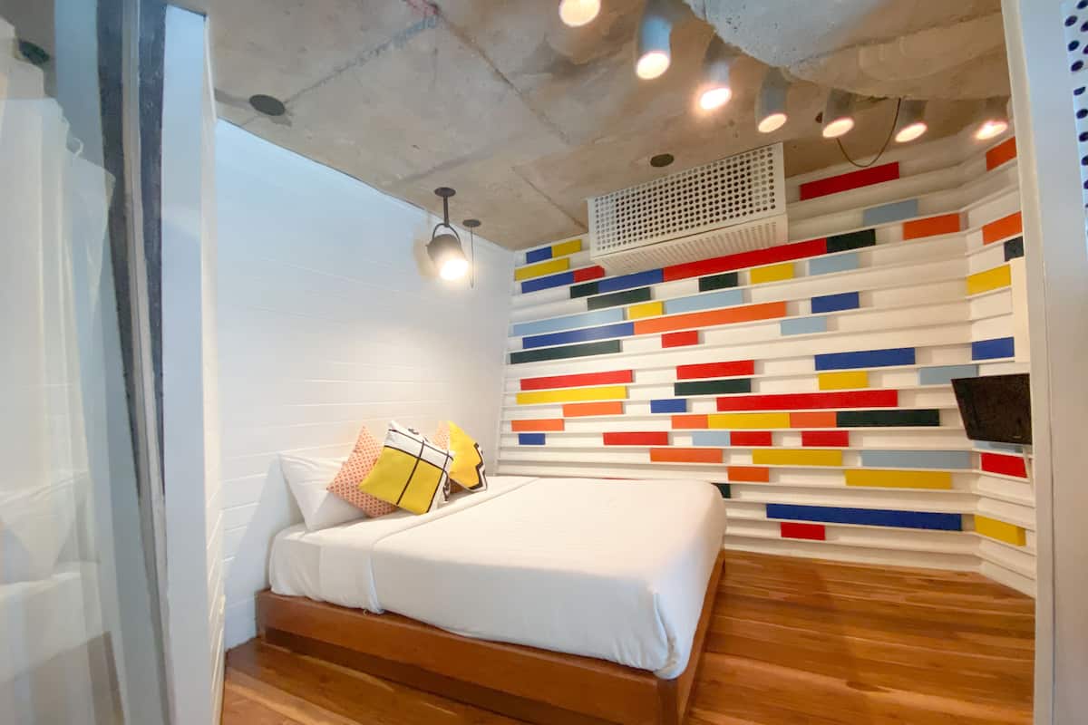 A modern bedroom with a wooden bed, white bedding, and a colorful, abstract wall design.