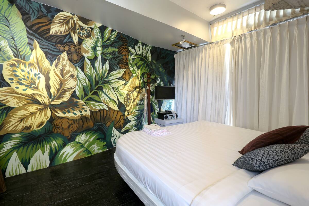A hotel room with a bed, white curtains, a flat-screen TV, and a wall mural of tropical leaves and a tiger.