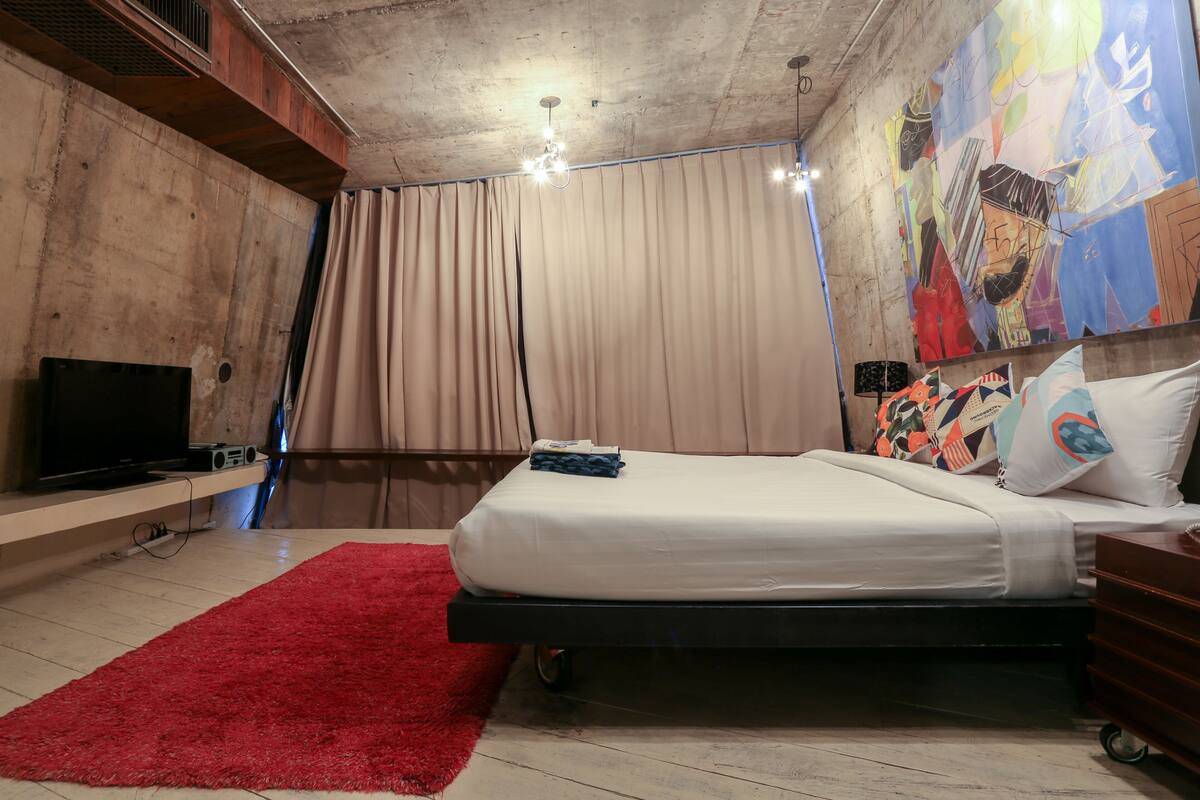 A bedroom with a bed, a TV, a red rug, and a large abstract painting on the wall.
