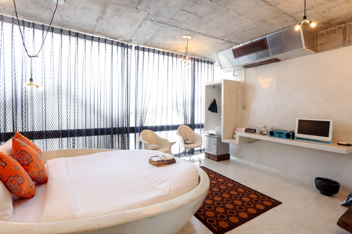 A modern hotel room with a large bed, a desk, a chair, a TV, and a window with sheer curtains.