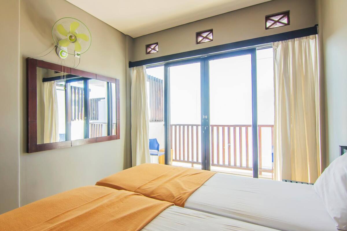 A hotel room with a bed, a ceiling fan, a large window with curtains, and a balcony with a railing.