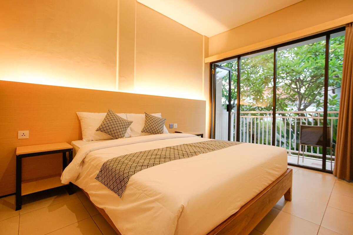 A bedroom with a large bed, a nightstand, and a view of a balcony with greenery.