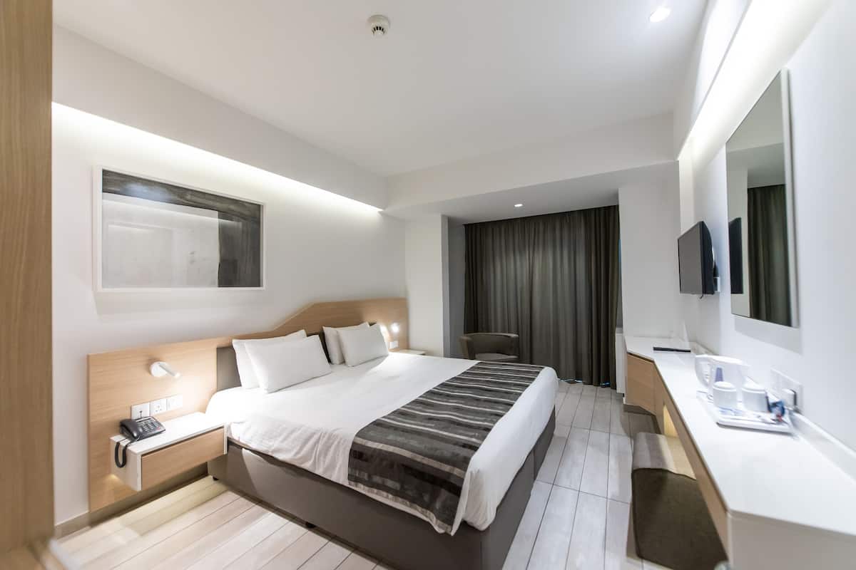 A modern hotel room with a large bed, bedside table with a phone, a desk with a mirror, and a TV.