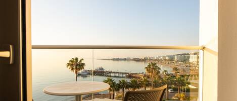 A balcony with a round table and a wicker chair, overlooking a coastal cityscape with a pier and palm trees.