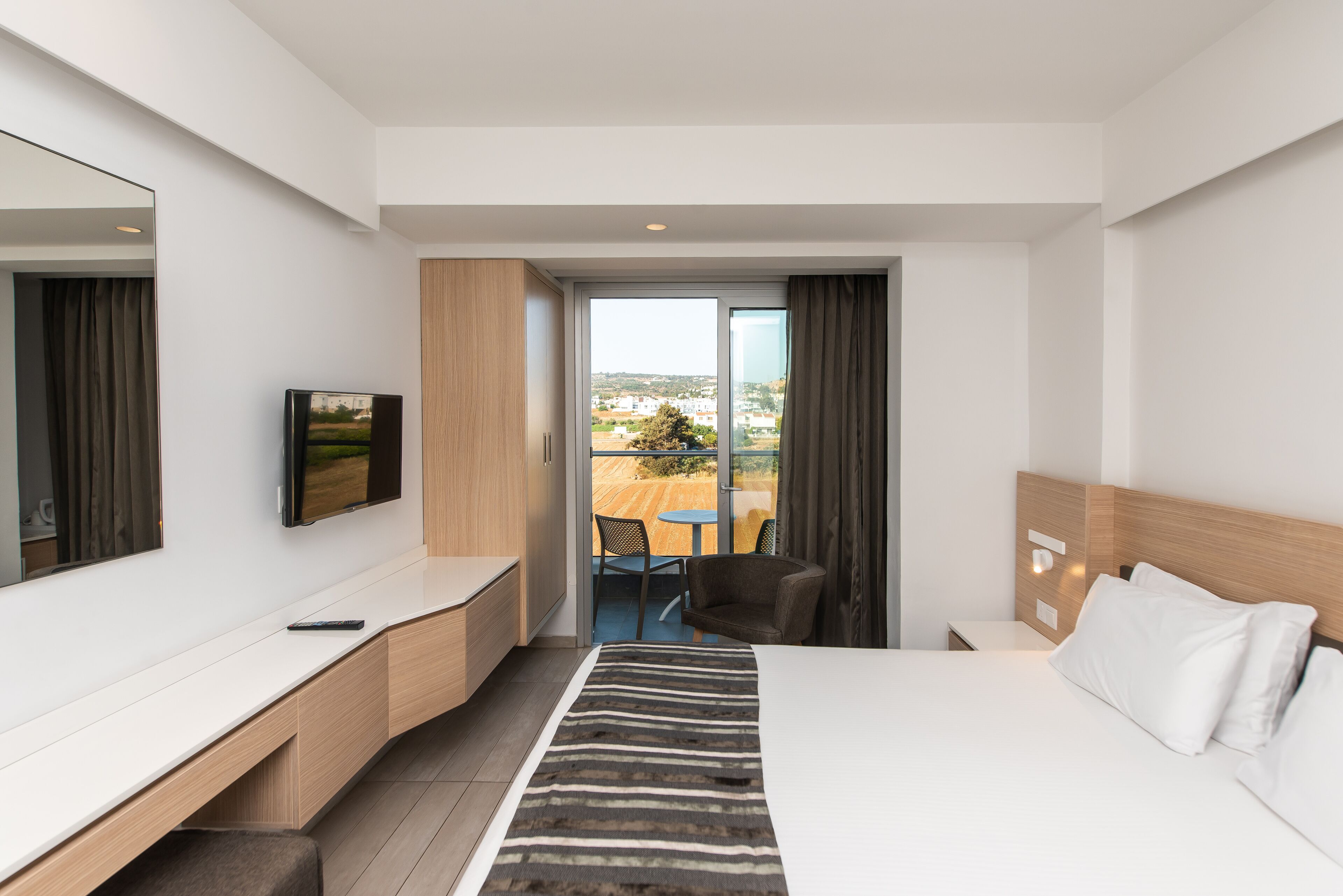 A modern hotel room with a large bed, a desk, a television, and a balcony with a table and chairs.