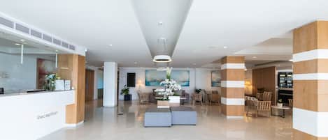 Uma área de recepção moderna com um balcão branco, um espaço de estar com sofás e cadeiras, e um quadro grande na parede.