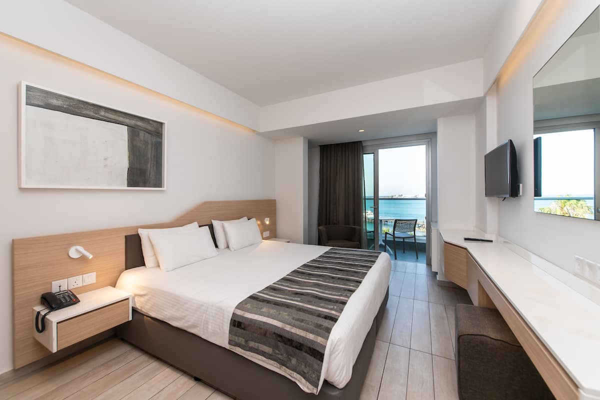 A modern hotel room with a large bed, bedside table with a phone, a flat-screen TV, and a view of the sea through a large window.
