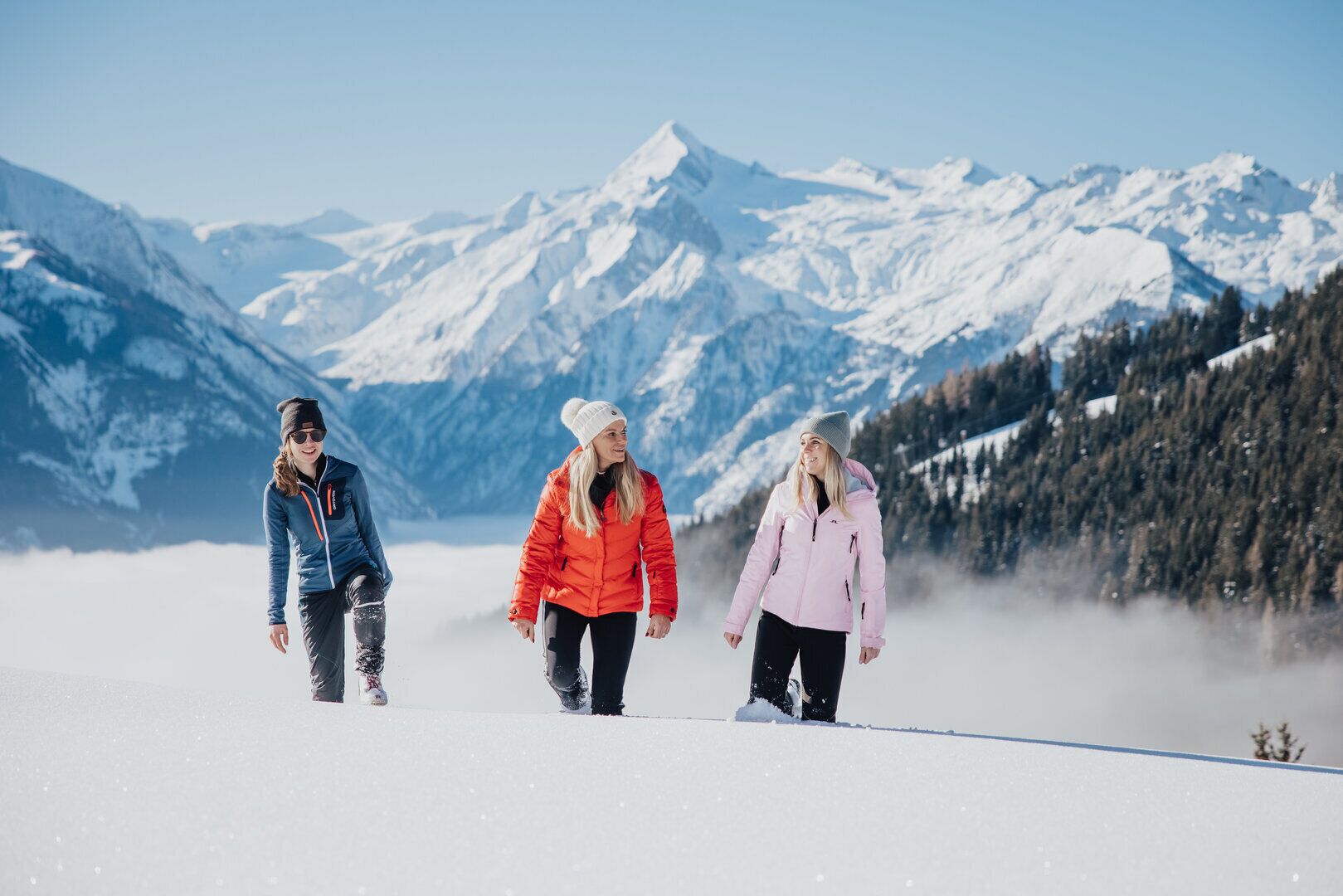 Tres personas caminando por un sendero cubierto de nieve, con montañas al fondo.
