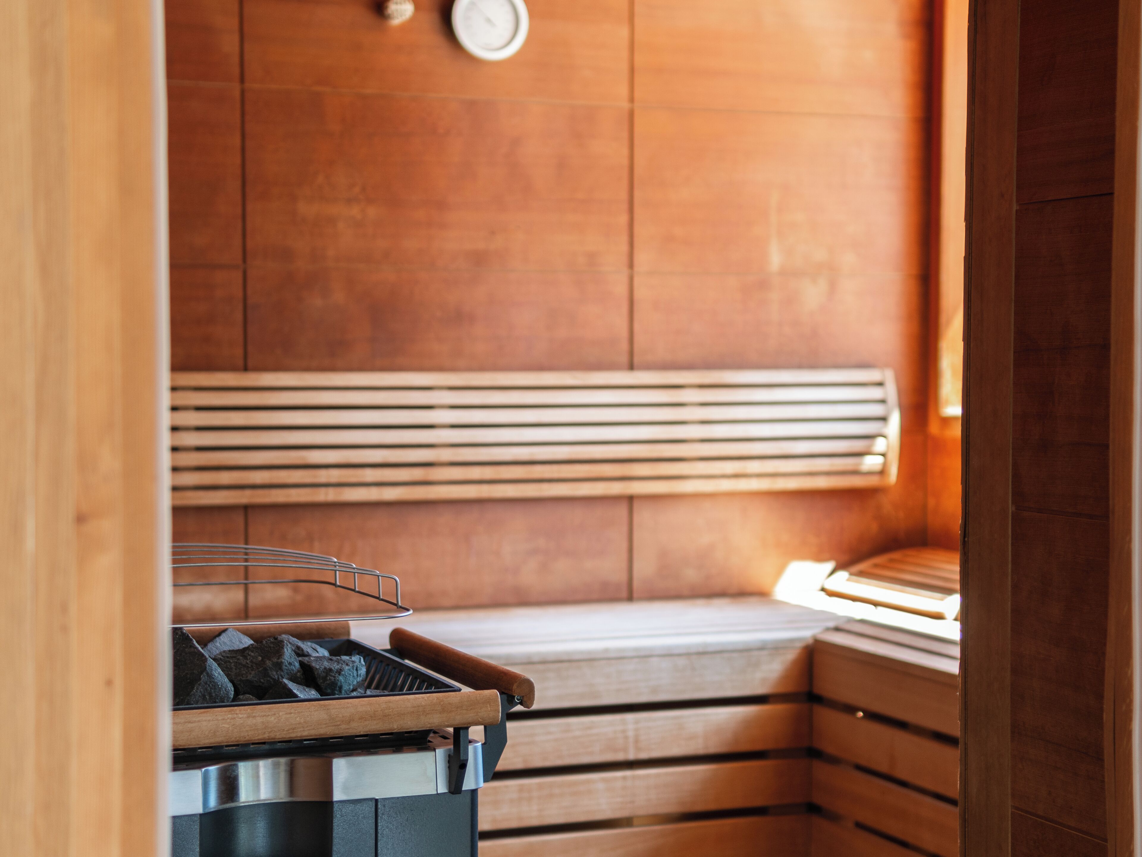 Sauna, jossa on puinen penkki, metallinen kannellinen astia ja seinälle kiinnitetty kello.