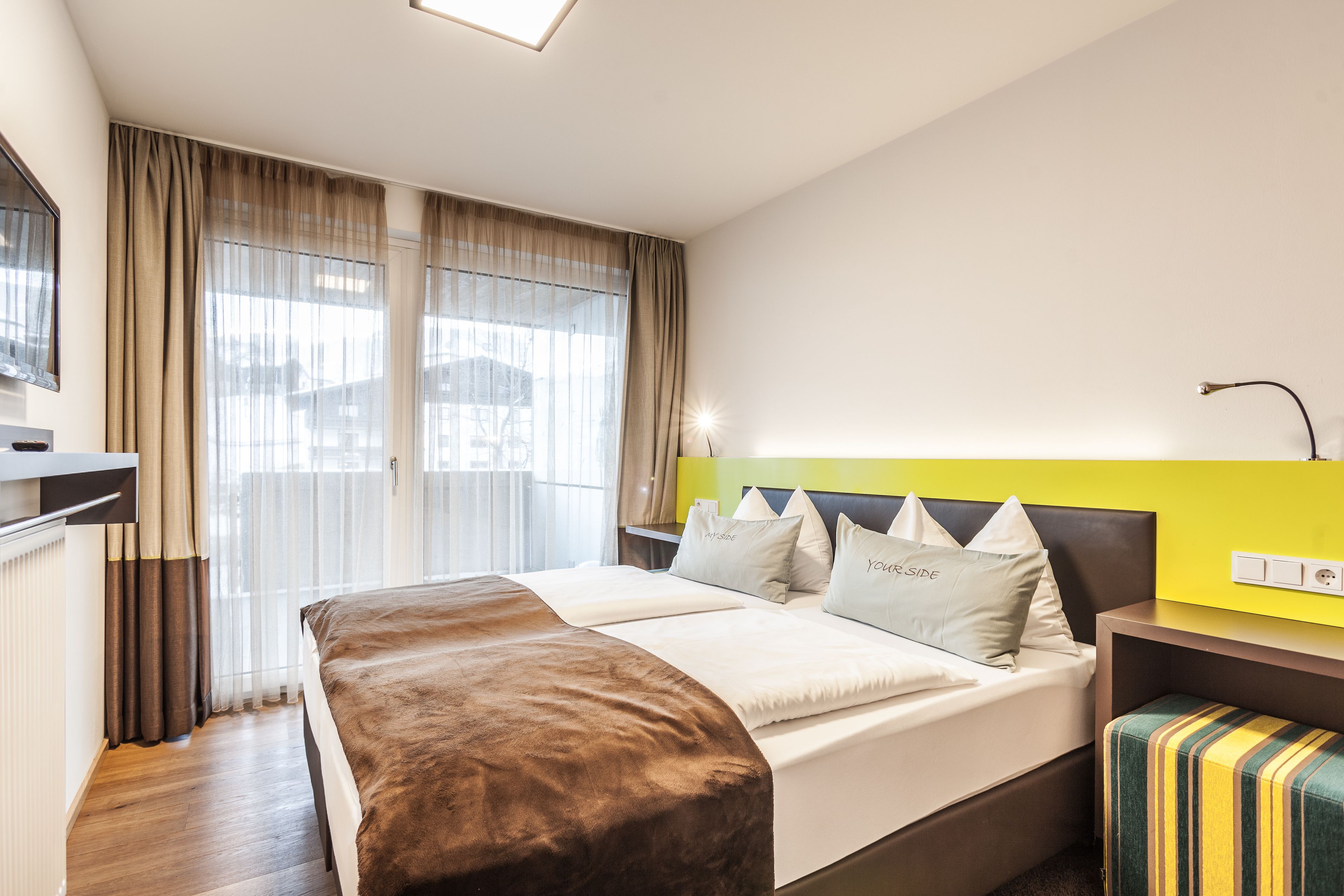 Une chambre d’hôtel moderne avec un grand lit, une table de chevet, un banc et une vue sur la ville à travers des rideaux transparents.