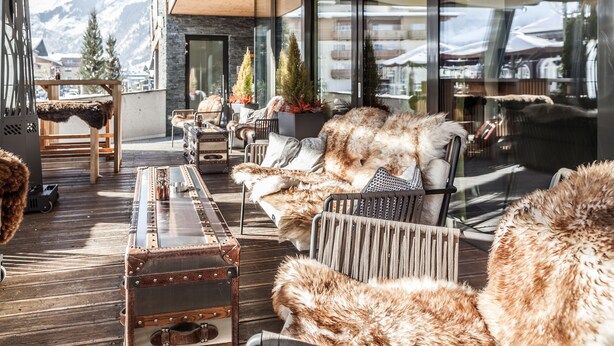 A cozy outdoor seating area with rustic furniture, a vintage suitcase, and a view of mountains through large windows. - Lederer Boutique Hotel  (Kaprun)