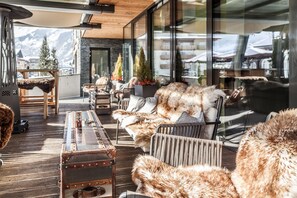 A cozy outdoor seating area with rustic furniture, a vintage suitcase, and a view of mountains through large windows. - Lederer Boutique Hotel  (Kaprun)