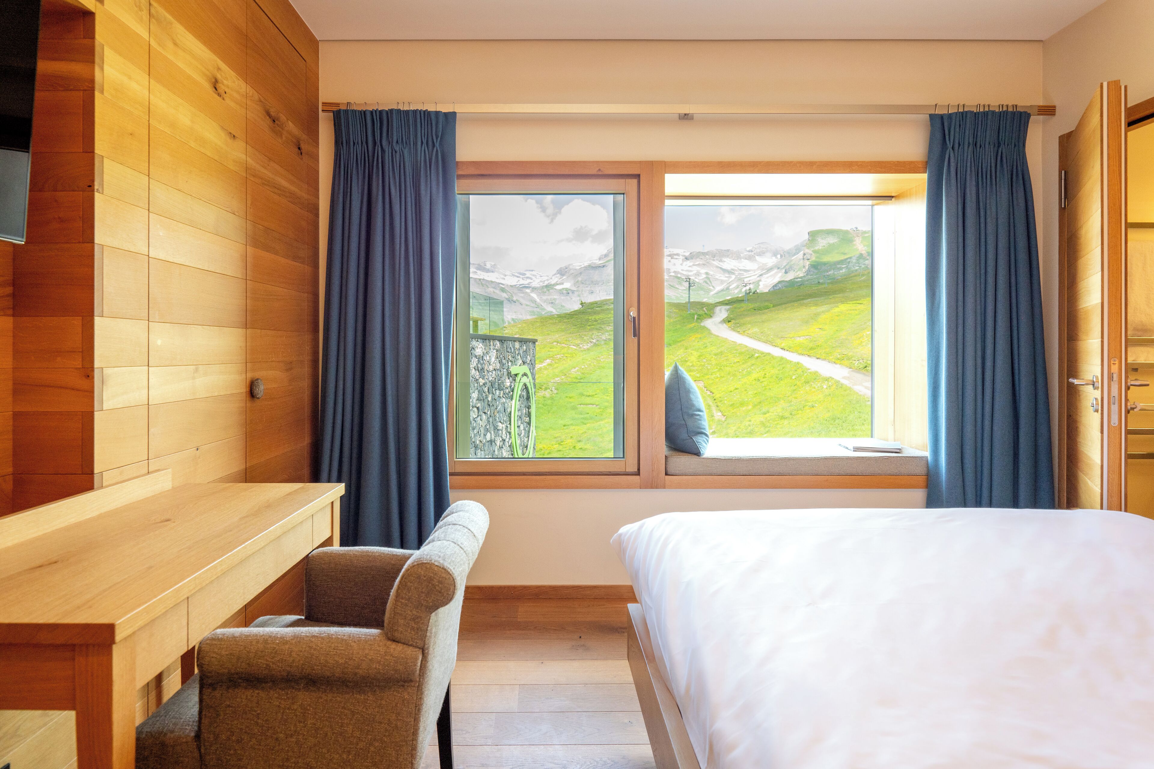 A bedroom with a wooden wall, a bed, a desk, and a window with a view of a mountain landscape.