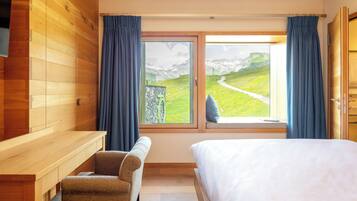 A bedroom with a wooden wall, a bed, a desk, and a window with a view of a mountain landscape.