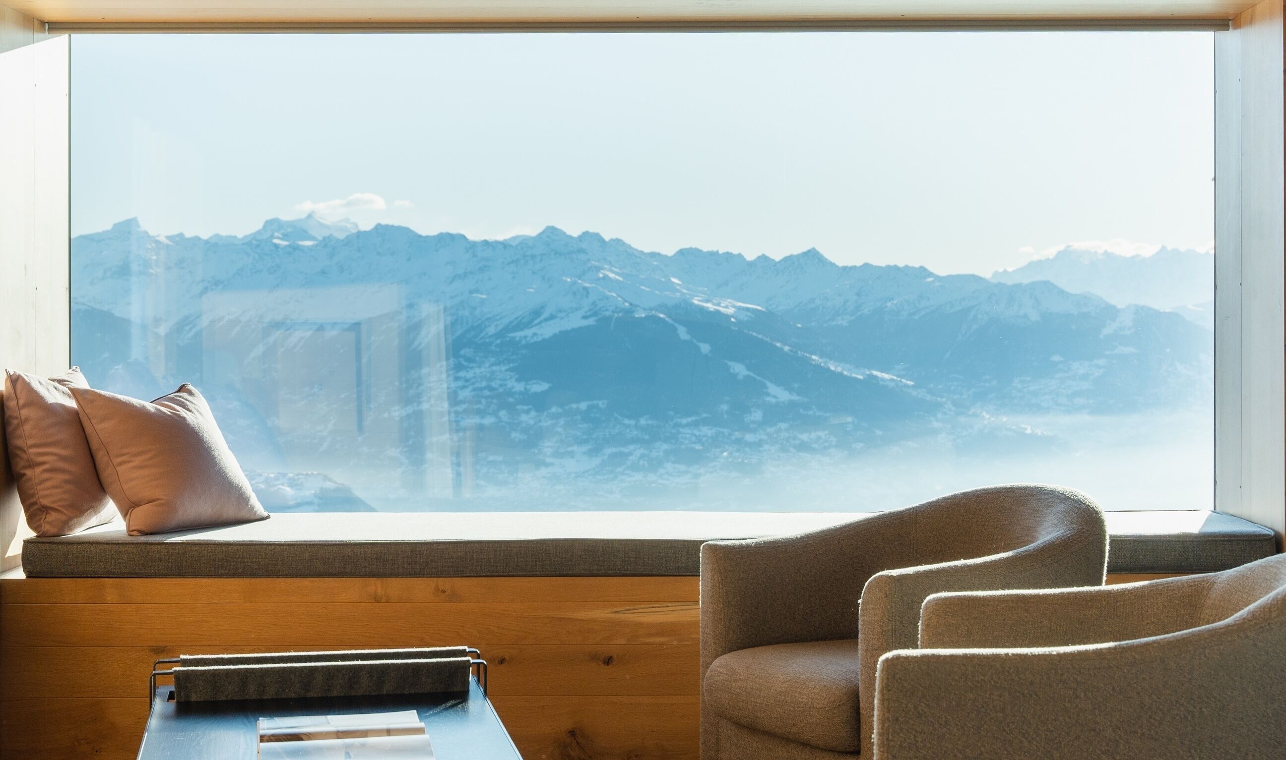 Habitación con un amplio ventanal con vistas a una cordillera. Hay un banco de madera con cojines y una pequeña mesa con una bandeja.