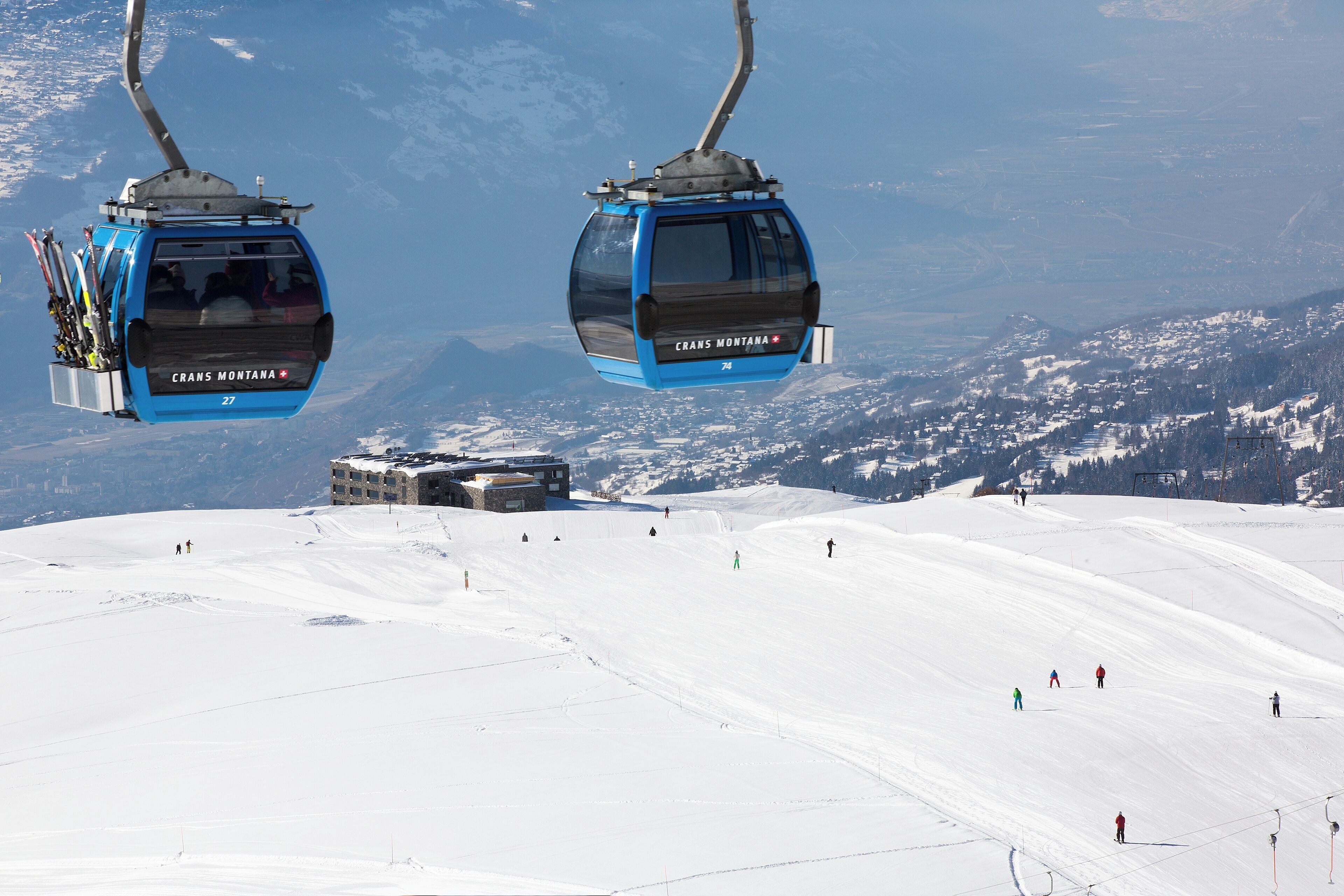 Eine Seilbahn mit Gondeln, beschriftet mit 'CRANS MONTANA', und ein schneebedeckter Hang mit Skifahrern.