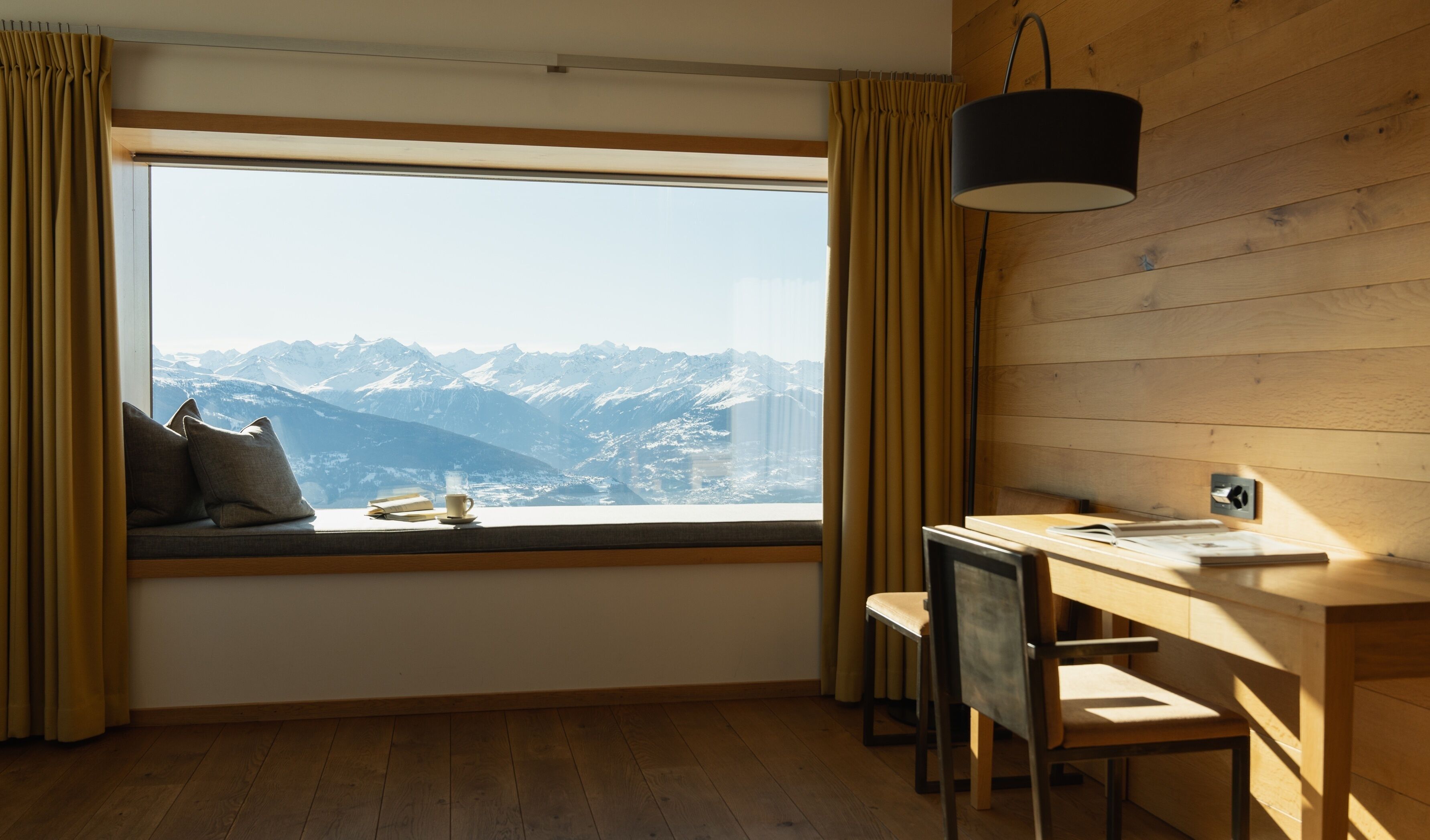 A room with a large window overlooking a mountainous landscape, a wooden desk with a chair, and a lamp.