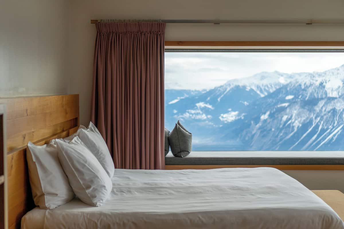 A hotel room with a bed, a window with a view of snow-capped mountains, and a wooden headboard.