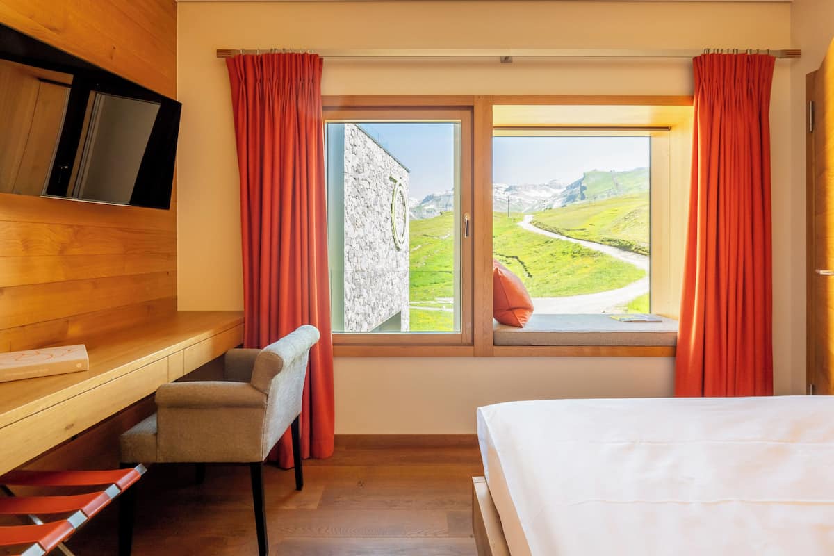 A bedroom with a bed, a desk with a chair, a TV, and a window with curtains.