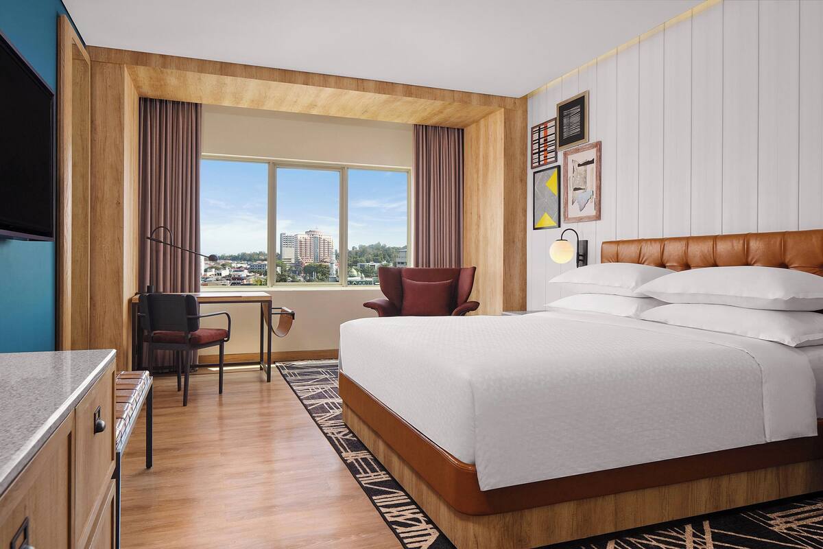 Une chambre d’hôtel avec un grand lit, un bureau, une chaise et une vue sur la ville grâce à de grandes fenêtres.