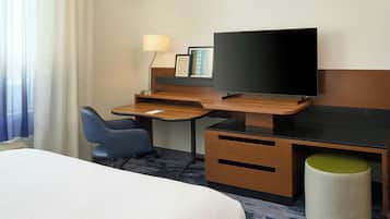 A hotel room with a bed, a desk with a chair, a television, a lamp, and a small stool.