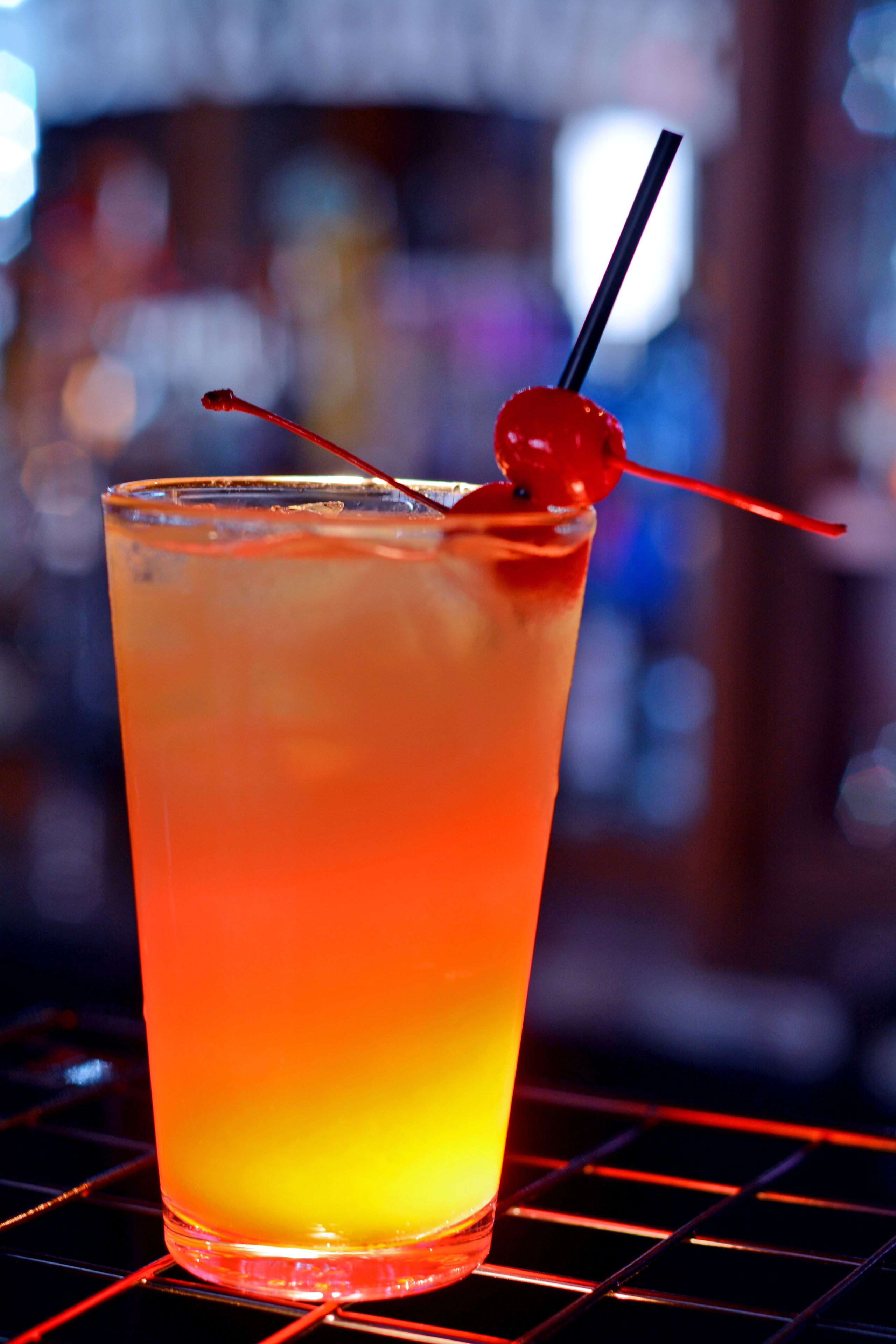 A tall glass with a gradient color from yellow to orange, a cherry on a straw, and a blurred background.