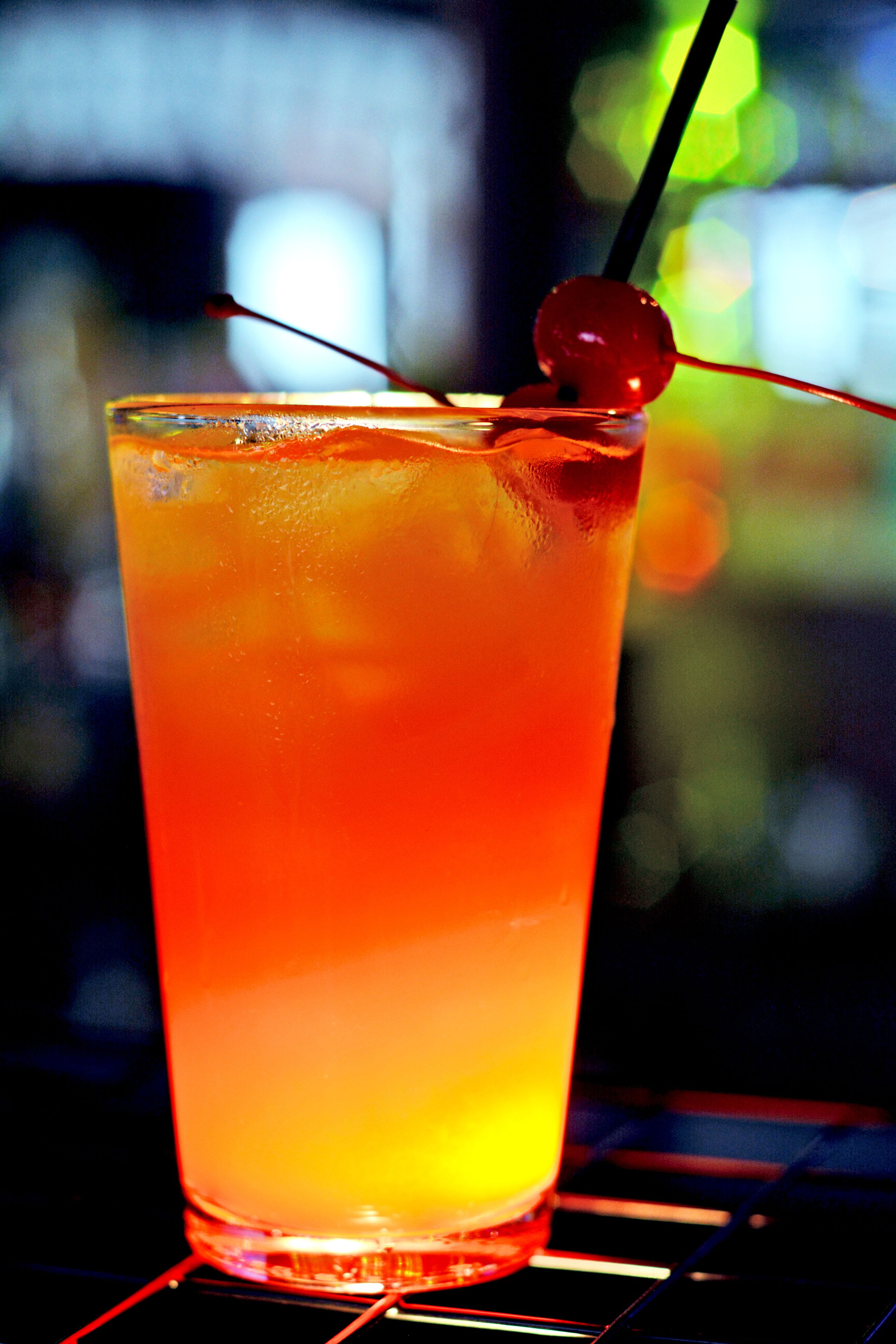 A tall glass with a gradient of orange and yellow liquid, a cherry on the rim, and a straw.