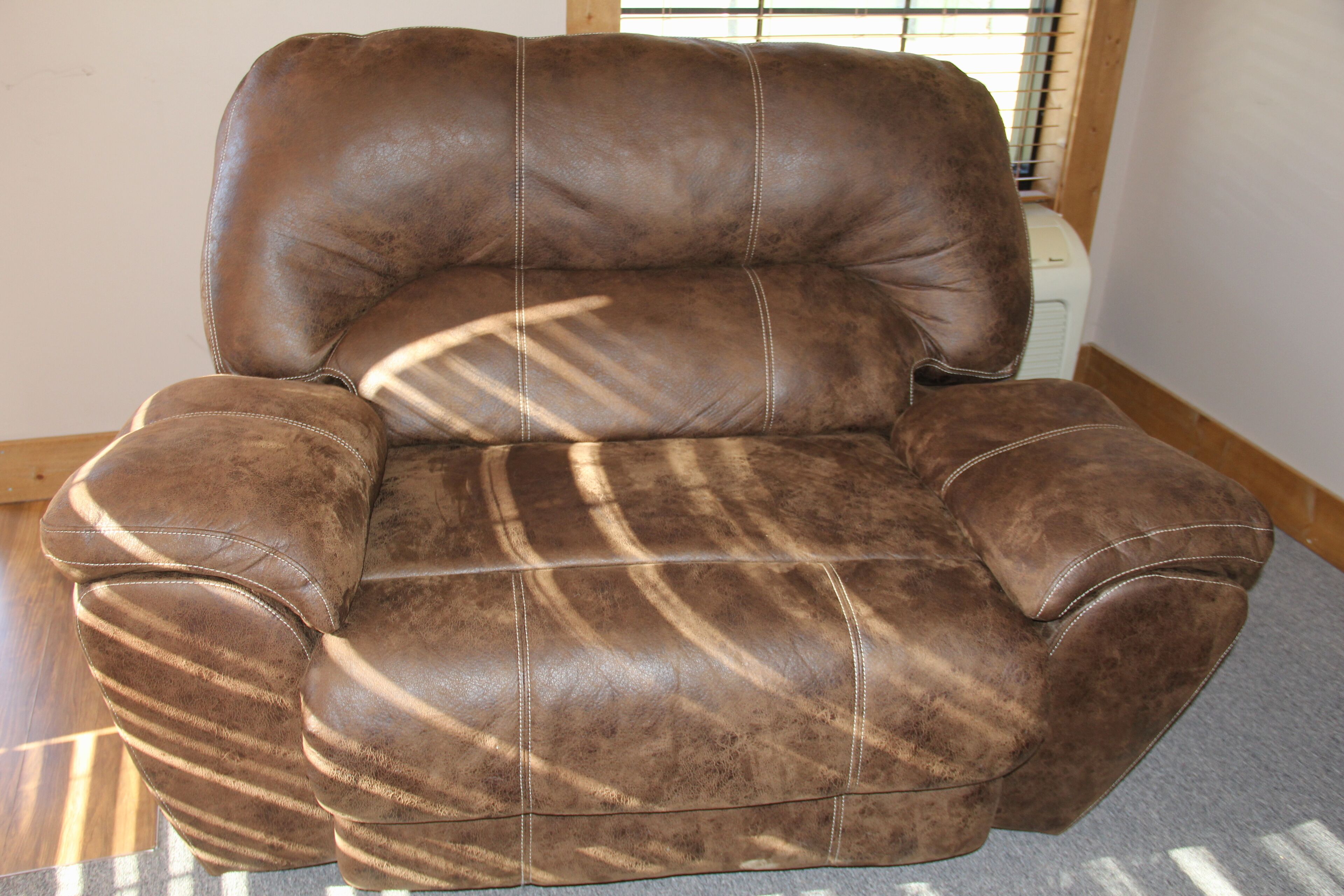 Un sillón de cuero marrón con un respaldo alto y reposabrazos, ubicado en una habitación con piso de madera y una ventana con persianas.