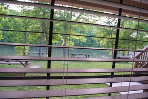 Vista a través de persianas de un espacio verde con mesas de picnic y una terraza de madera.