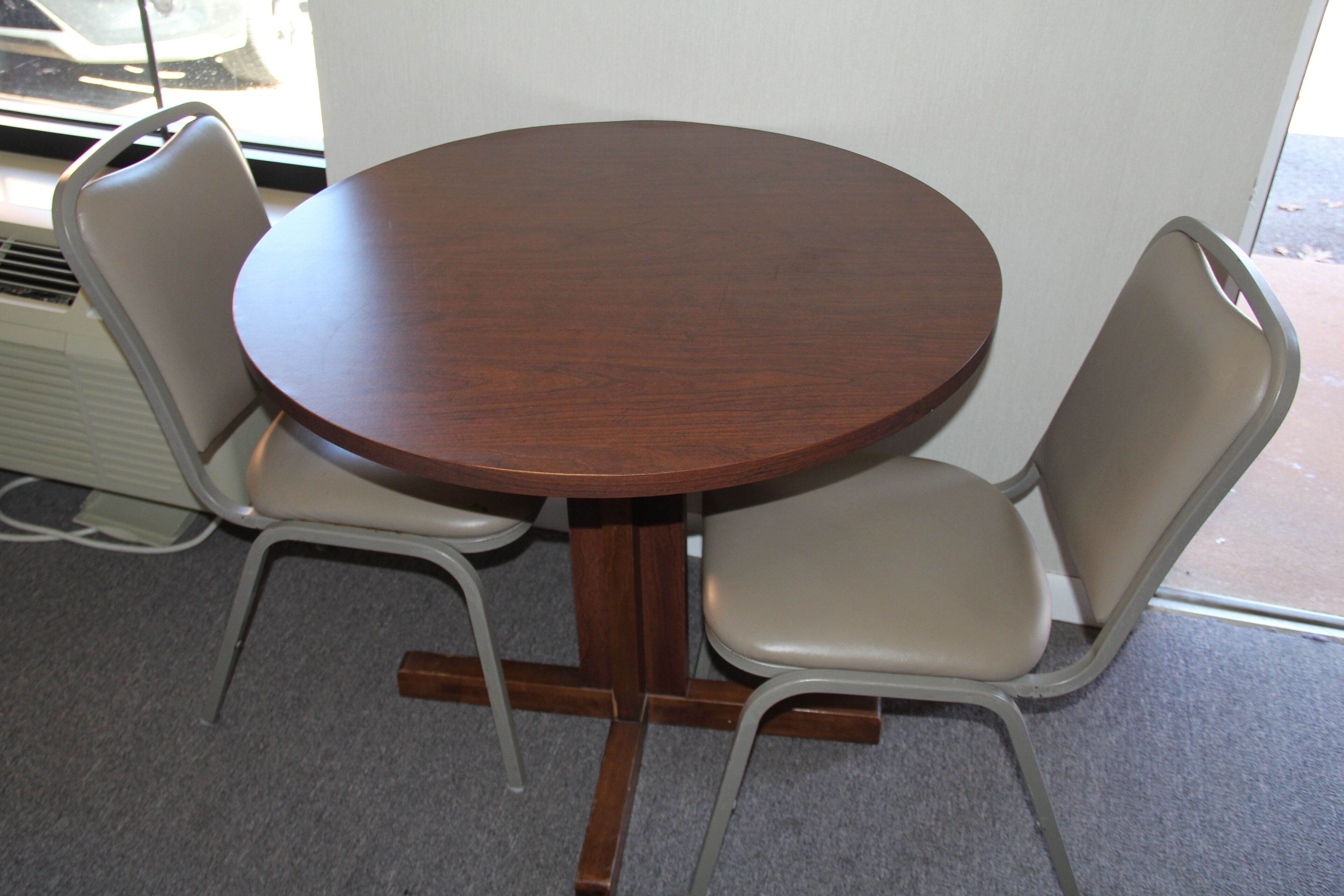 A round wooden table with four chairs, a white radiator, and a window with a view of the street.