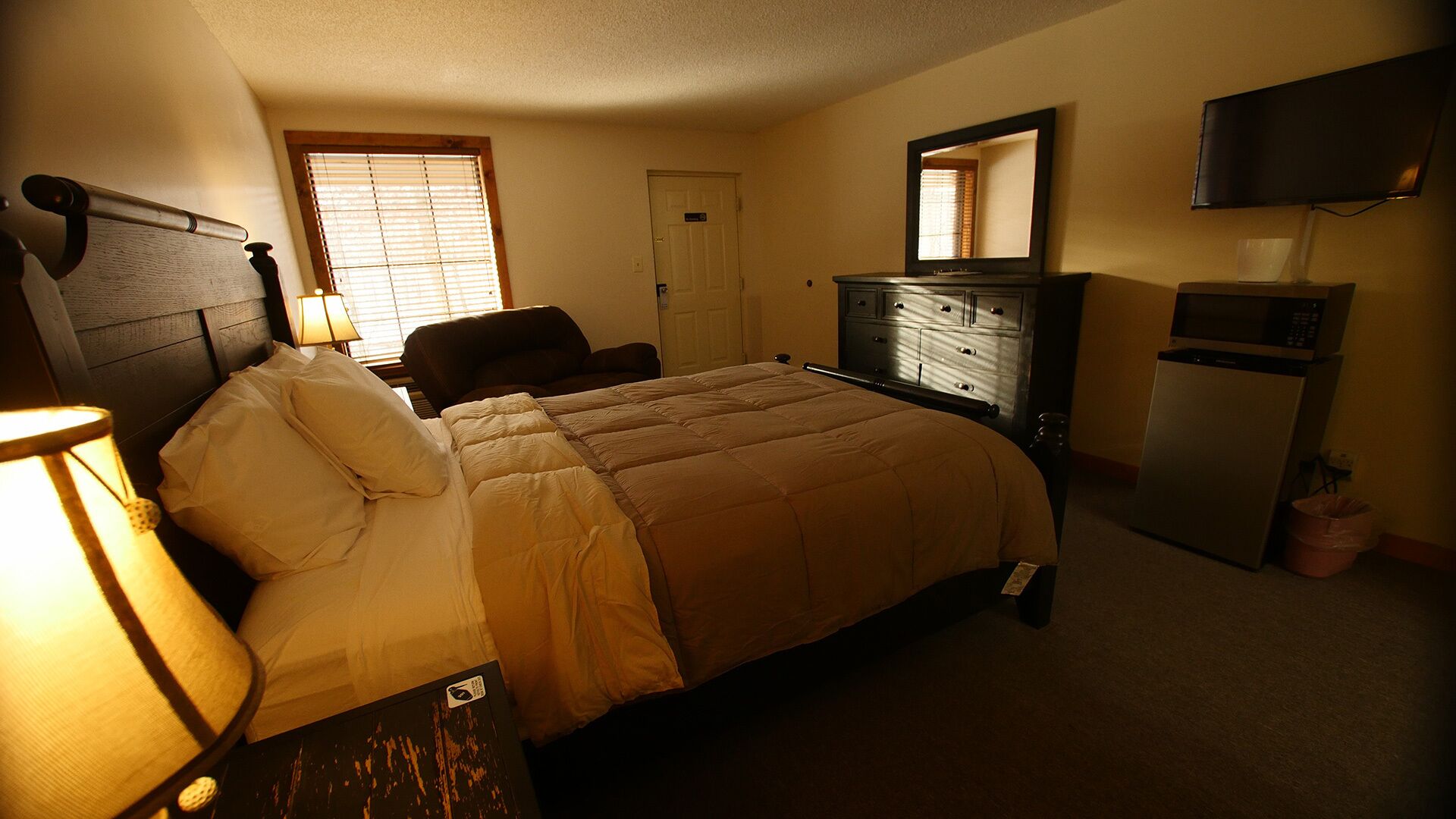 A bedroom with a bed, a dresser, a chair, a desk with a lamp, a television, and a window with blinds.