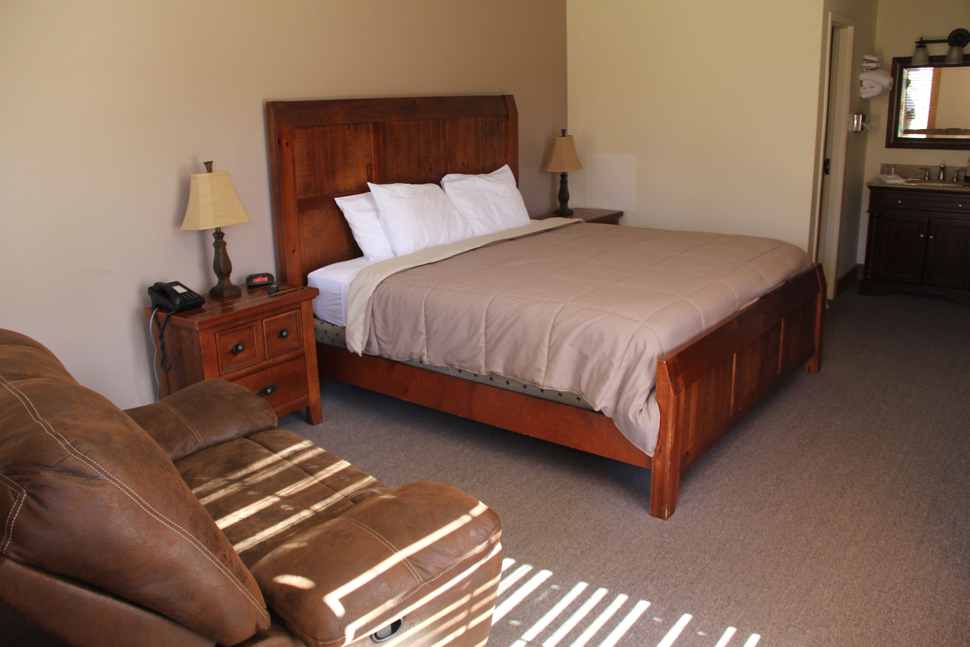 Un dormitorio con una cama de madera, dos mesitas de noche con lámparas, un sofá de cuero marrón y un baño visible a través de una puerta abierta.
