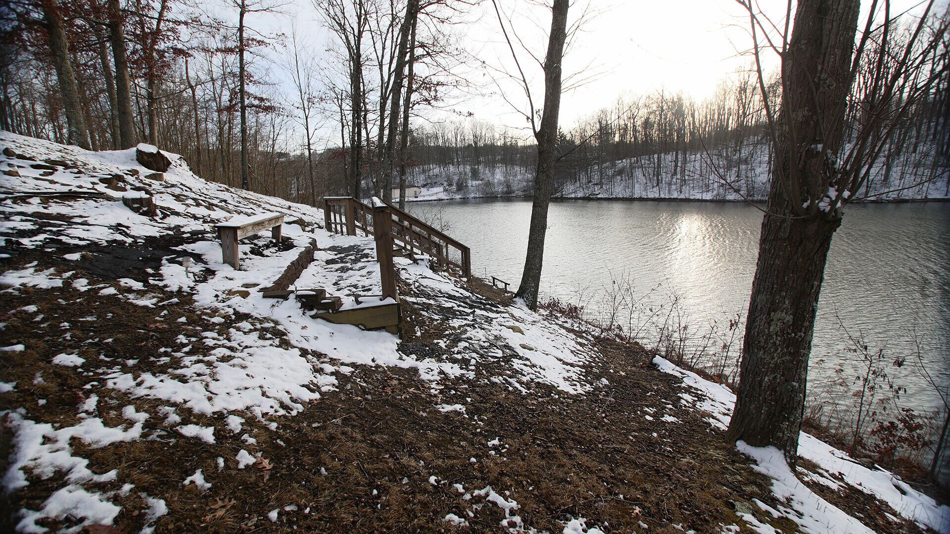 A snow-covered outdoor area with a wooden staircase leading down to a body of water, surrounded by trees.