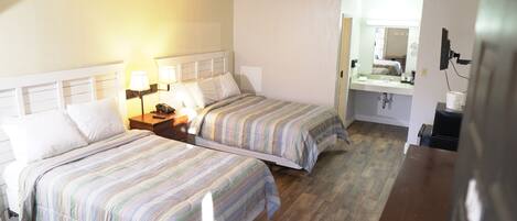 A hotel room with two beds, a nightstand with a lamp, a small table, a bathroom with a sink and mirror, and a television.