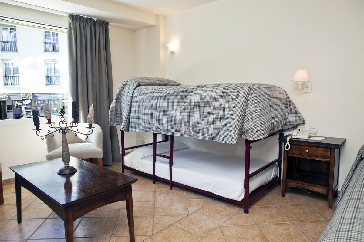 A room with a bunk bed, a bedside table with a lamp, a small nightstand, and a window with curtains.