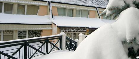 A hotel building with a sign reading "Hotel Telecaba" covered in snow, a black metal railing, and snow-covered trees.